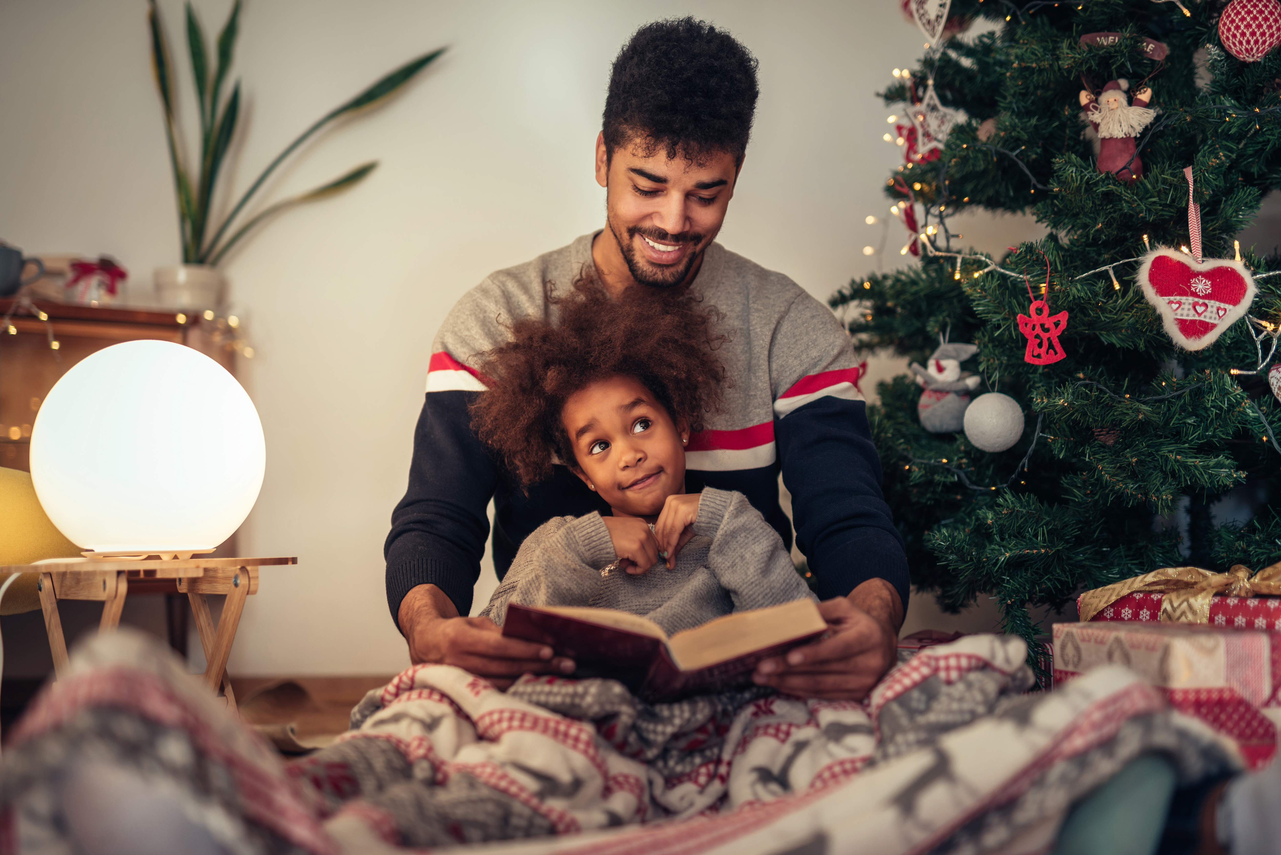 Father and son reading a book sitting by a Christmas tree. Son sits on father's lap, both are wearing pajamas. 